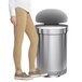 The lower half of a person standing next to a simplehuman brushed stainless steel semi-round step-on trash can.