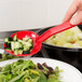 A hand holding a Thunder Group red polycarbonate salad bar spoon with cucumbers and lettuce over a plate of salad.