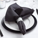A folded black Intedge cloth napkin on a white plate with silverware.