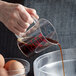 A hand pouring brown liquid from a glass jar into a Carlisle clear polycarbonate measuring cup.