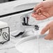 A person's hand pressing the Waterloo deck-mounted faucet to pour water into a sink.