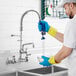 A man wearing blue and yellow gloves using a Waterloo wall-mounted pre-rinse faucet to spray a sink in a professional kitchen.
