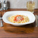 A CAC square ivory china bowl filled with spaghetti, sauce, and parmesan cheese on a table.