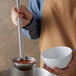 A person using a Vollrath stainless steel ladle to pour liquid into a white bowl.
