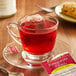 A glass cup of Bigelow Pomegranate Pizzazz herbal tea with a tea bag on a saucer.