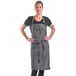 A woman wearing a Mercer Culinary black denim bib apron with a leather towel holder over a black shirt.