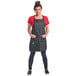 A woman wearing a Mercer Culinary black denim bib apron with brown leather straps.