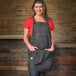 A woman wearing a black denim Mercer Culinary apron with brown leather straps.
