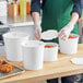 A person in a green shirt wearing a plastic glove puts food in a white Choice food bucket with a lid.