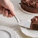 A hand using a Tablecraft stainless steel pastry server to cut a piece of chocolate cake on a plate.