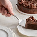A hand using a Tablecraft stainless steel pastry server to cut a piece of chocolate cake.