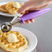 A hand using a purple Hamilton Beach orchid thumb press scooper to add a scoop of ice cream to a waffle.