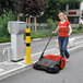A woman in a red shirt pushing a Bissell Commercial outdoor power sweeper on a sidewalk.