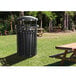 An Ex-Cell Kaiser black round outdoor trash can with canopy on a grass field next to a wooden picnic table.