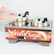 A person holding a Vollrath Country Kitchen soup warmer base with a container of soup on a counter.