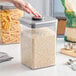 A hand using a measuring spoon to fill an OXO Good Grips food storage container with white grains.