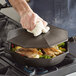 A hand using a white towel to clean a FINEX cast iron cover over food in a pan.