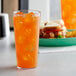 A Carlisle clear plastic tumbler filled with orange juice and ice on a table with a sandwich.