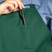 A person holding a pen in a Hunter Green Uncommon Chef bib apron with three pockets.