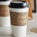 A 10-24 oz. natural Kraft coffee cup sleeve with a brown label on a table with two paper coffee cups.