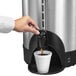 A hand pouring coffee from a Hamilton Beach stainless steel coffee urn.