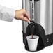 A hand pouring coffee from a Hamilton Beach stainless steel coffee urn into a cup.