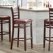 Three Lancaster Table & Seating mahogany wood ladder back bar stools with burgundy vinyl seats at a wooden bar.