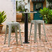 A black Lancaster Table & Seating Excalibur outdoor table base column with white top and gray stools on a brick patio.
