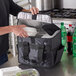 A hand placing a white container of food inside a black American Metalcraft insulated delivery bag.