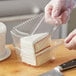 A hand wearing a plastic glove using a knife to cut a piece of cake into a clear plastic container with a high dome lid.