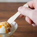 A hand holding a Royal Paper wooden taster spoon over a scoop of ice cream.