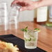 A person pouring clear liquid into a Duralex glass filled with ice and mint on a black tray.