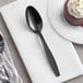 A Visions black plastic teaspoon wrapped in plastic on a white napkin next to a chocolate cake.