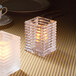 A Hollowick clear glass square candle holder with a lit candle on a table.