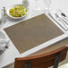 A beige Front of the House Rattan woven vinyl placemat on a table with a glass of wine and silverware.
