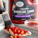 A hand using a paint brush to apply Sauce Craft Sweet and Smoky BBQ Sauce to food on a grill.