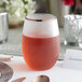 A Visions clear plastic stemless wine glass with a copper rim filled with red liquid on a table.