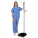 A woman wearing blue scrubs and pants standing next to a Cardinal Detecto digital scale.