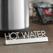 An American Metalcraft stainless steel tabletop sign with "Hot Water" on a table next to a kettle.