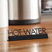 An American Metalcraft black laser-cut table sign that says "Hot Water" on a counter.