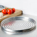 An American Metalcraft standard weight aluminum pizza pan with perforations on a wooden surface with tomatoes and cheese.