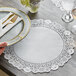 A hand holding a Hoffmaster lace doily under a plate with a fork and knife on a table.