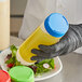A hand in a black glove holding a Tablecraft bottle with a yellow liquid and a blue lid.