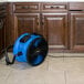A blue and black XPOWER FC-300 shop fan on a tile floor next to wood cabinets.