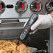 A hand holding a Taylor digital infrared thermometer with a folding thermocouple probe over a professional kitchen counter.