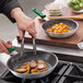 A person using a green Choice silicone handle sleeve on a pan with sausages and potatoes.
