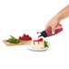 A hand holding a clear OXO squeeze bottle pouring red sauce onto a piece of cheesecake.
