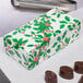 A white Holly candy box with green leaves and red berries on a counter next to chocolates.