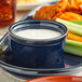 A blue Acopa stoneware ramekin filled with white liquid on a plate with celery and carrots.