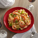 A Tuxton Concentrix cayenne china plate with pasta, shrimp, and basil on a table with a fork and a glass of wine.